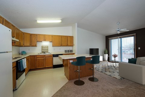 a kitchen with white appliances and wooden cabinets. Theres a living room in the background with a gray accent wall. Fargo, ND Bridgeport Apartments