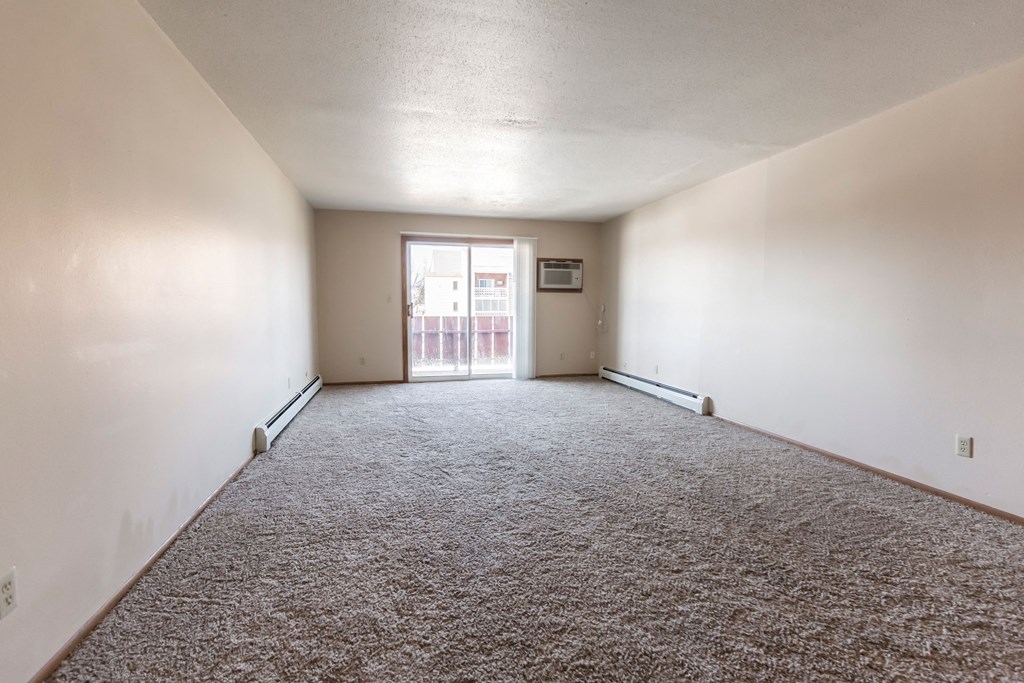 Grand Forks, ND Cherry Creek Apartments. A vacant living room in the unit 223 with a glass sliding door and white walls with a carpeted floor.
