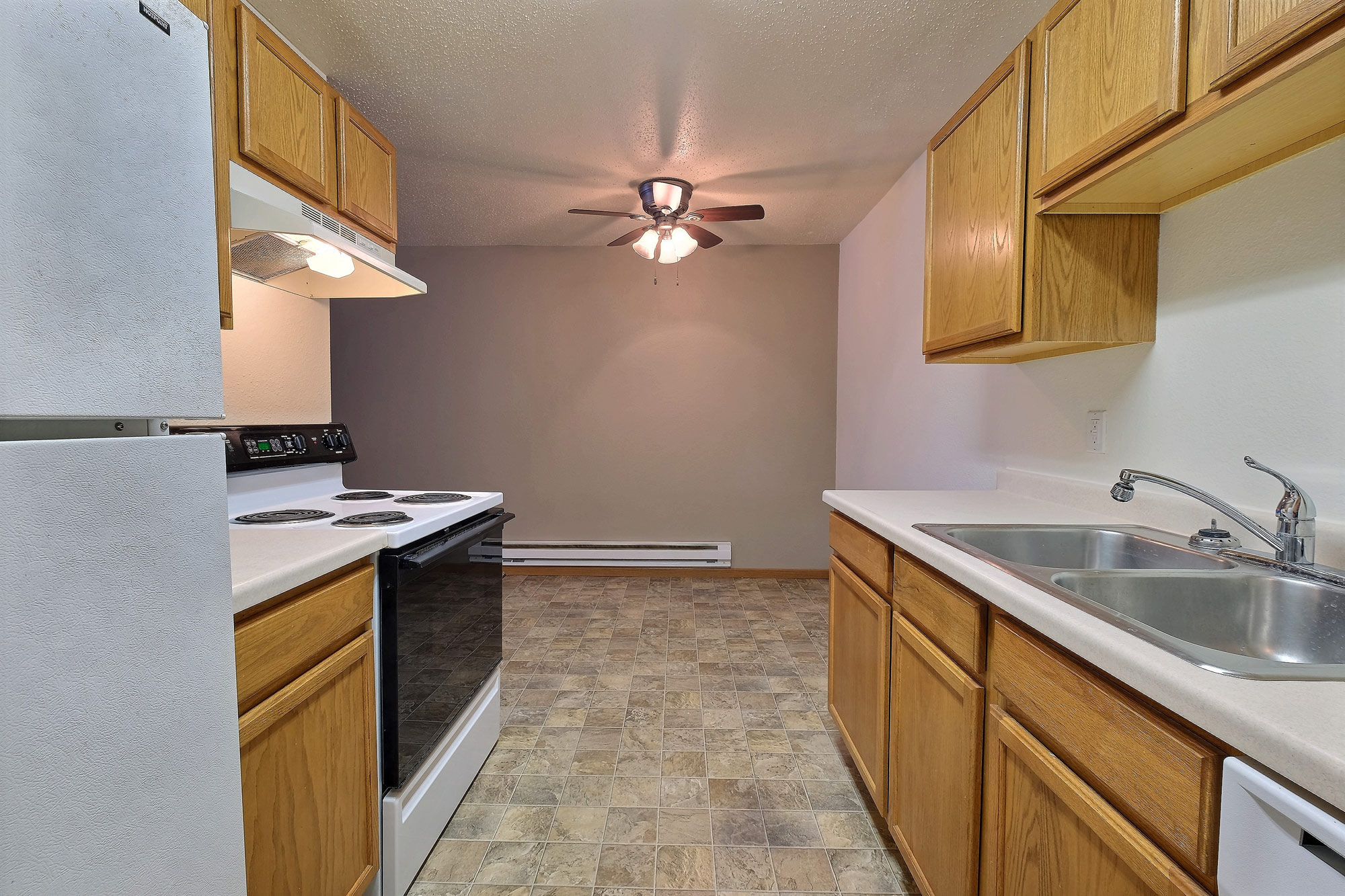 a kitchen with white appliances and a dining room in the background with a ceiling fan. Fargo, ND Dakota Manor Apartments
