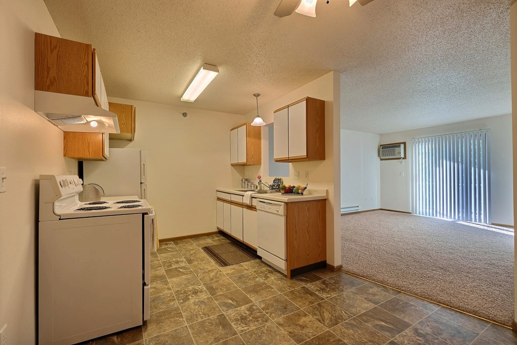 A kitchen with white cabinets and white appliances with a living room with a door to a balcony. Fargo, ND Eagle Run Apartments.
