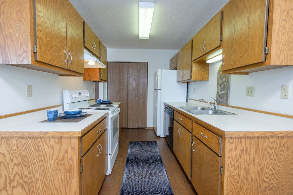 a kitchen with wood cabinets and white appliances and a blue rug. Fargo, ND Park Circle Apartments.