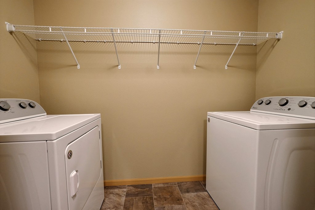 two washers and a dryer in a laundry room with a rack on top. Fargo, ND Urban Plains Apartments