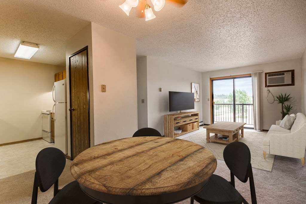 Bismarck, ND Eagle Sky I Apartments | Dining Bismarck, ND Eagle Sky I. A dining room with a four chair table. a living room with a couch, coffee table, and a flat screen tv in the background. The glass sliding door in the living room brightens the room with natural light.