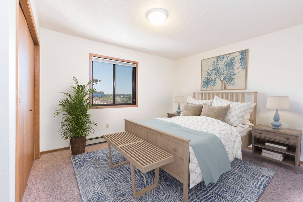 Bismarck, ND Fairview Apartments. A bedroom that showcases a comfortable bed with neatly arranged bedding, a stylish bed bench, and a window that fills the space with natural light. It's a serene and inviting setting for rest and relaxation.