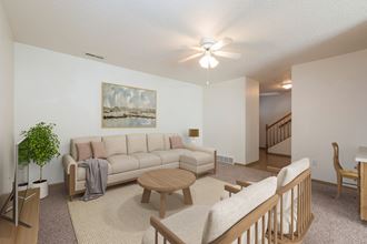 Bismarck Pebble Creek Apartments. The spacious living room is tastefully decorated with stylish furniture, ample natural light, and a cozy atmosphere. There's overhead lighting with a touch of natural light from the dining room. Access to the upstairs is in the background