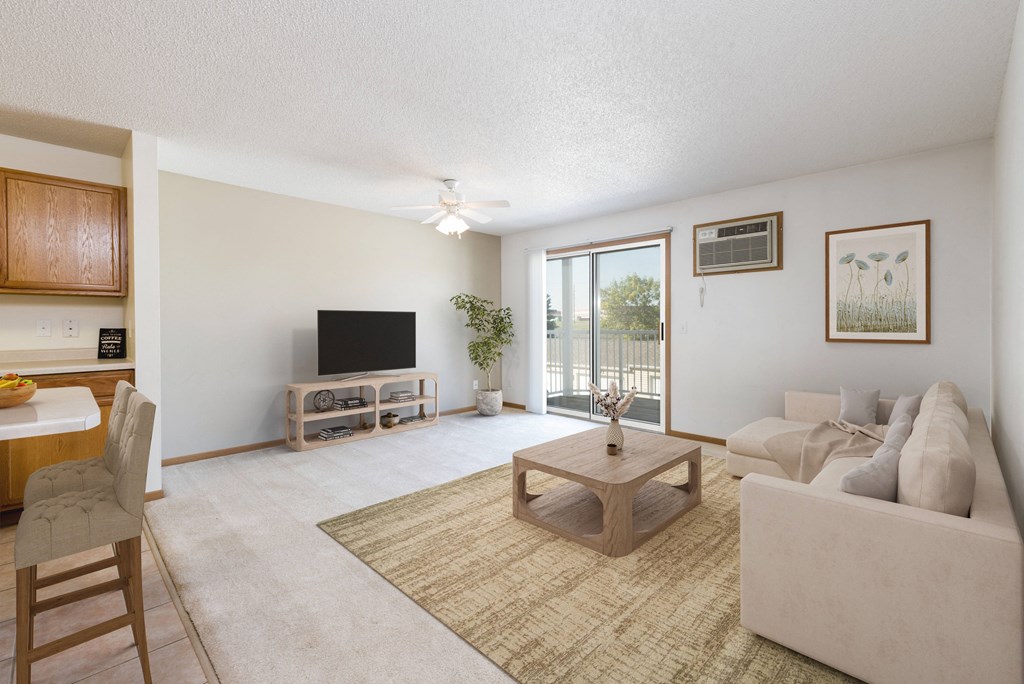 Sierra Ridge Apartments in Bismarck, ND | a living room with white walls and a sliding glass door. Theres a white couch, coffee table, tv and bar stools at the counter in the dining area.