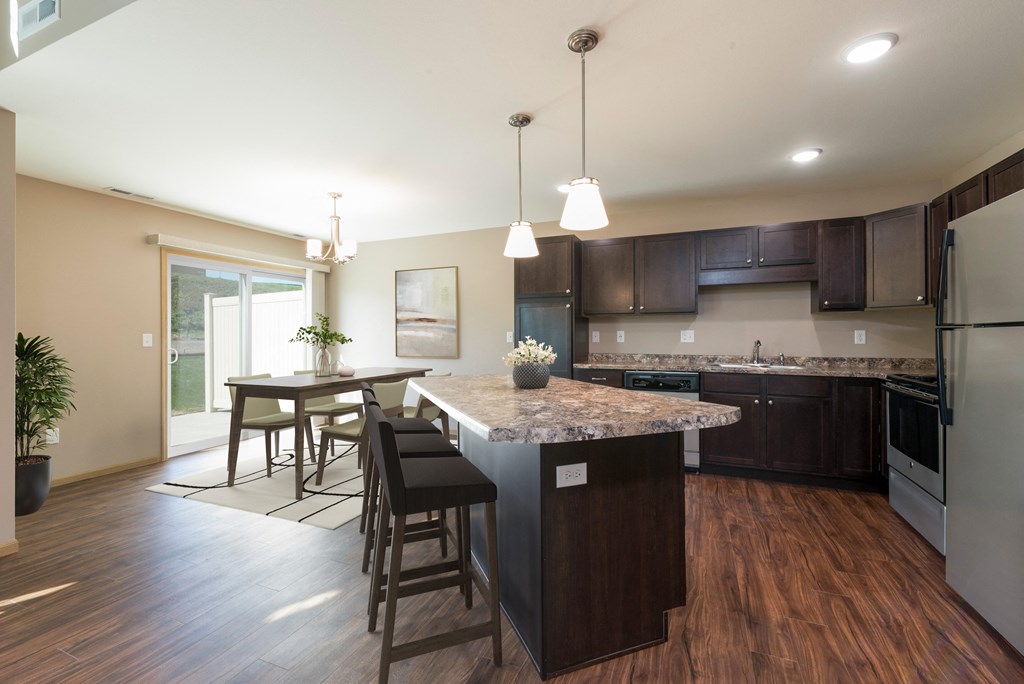 Bismarck, ND Stonefield Townhomes | Kitchen Bismarck, ND Stonefield Townhomes. A kitchen with dark wood cabinets and a large island with a granite countertop. The space features a well-appointed dining table, modern kitchen appliances, and tasteful decor. Bright natural light fills the room from the glass sliding door.