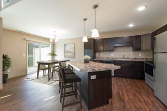 Bismarck, ND Stonefield Townhomes. A kitchen with dark wood cabinets and a large island with a granite countertop. The space features a well-appointed dining table, modern kitchen appliances, and tasteful decor. Bright natural light fills the room from the glass sliding door.