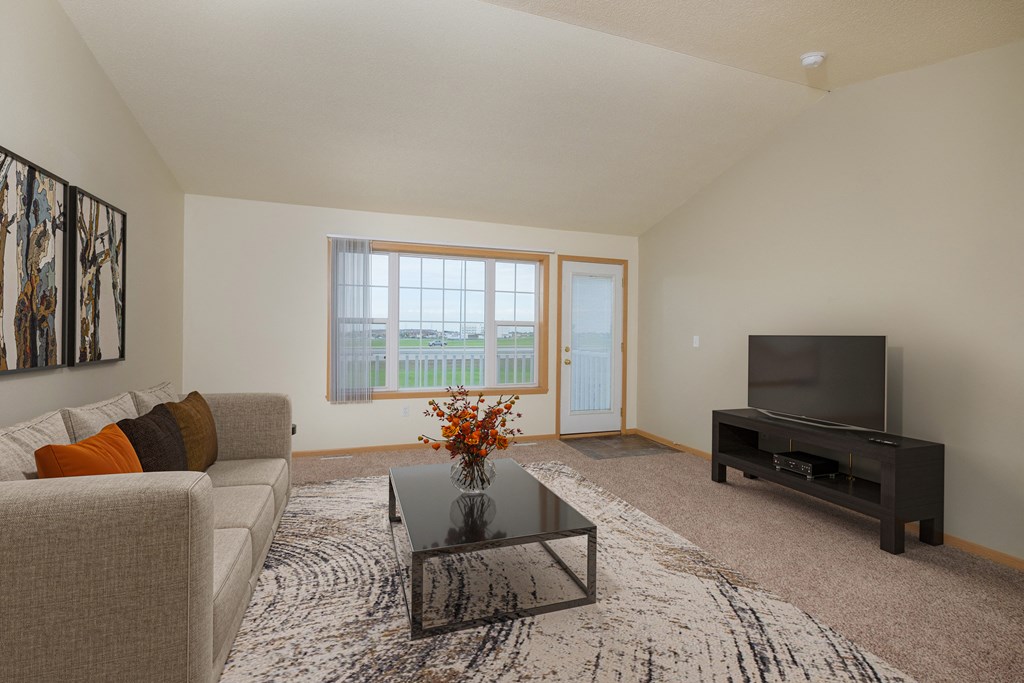 A living room with a couch, coffee table, and tv with an entry door and a window. West Fargo, ND Charleswood Townhomes.