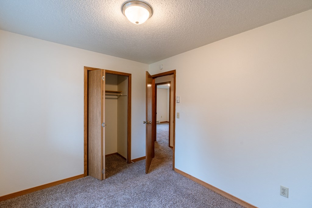 a bedroom with a carpeted floor and a hallway with two doors and a closet. Fargo, ND Galleria 3 Apartments