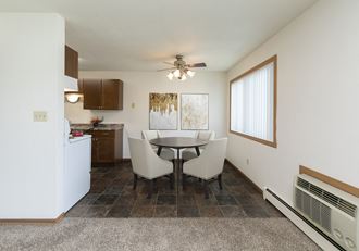 Fargo, ND Islander Apartments. A dining area with a four chair table that has a cailing fan with a light above it. A window brightens the room with natural light. Theres also a kitchen in the background