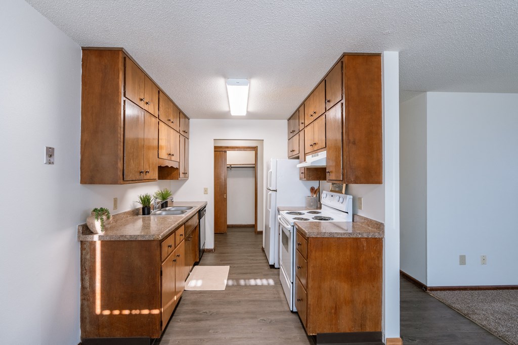 an open kitchen with wood cabinets and white appliances and a hallway with a door to. Fargo, ND Martha Alice Apartments