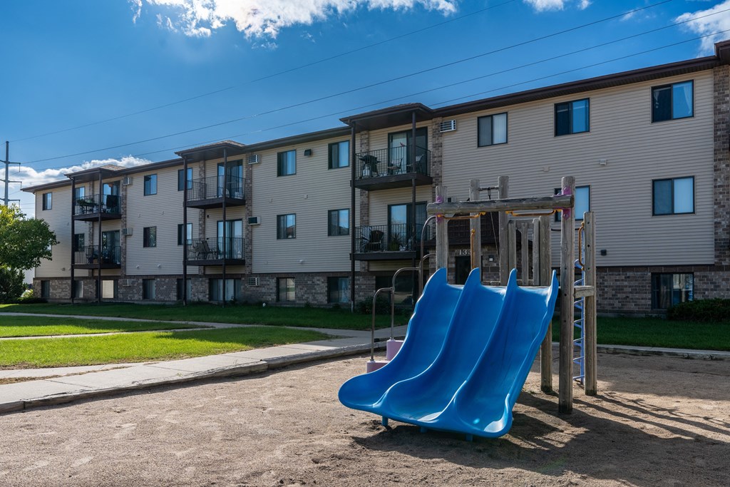 Fargo, ND Prairie Park Apartments | Playground Fargo, ND Prairie Park Apartments. A playground with two blue slides in front of an apartment building