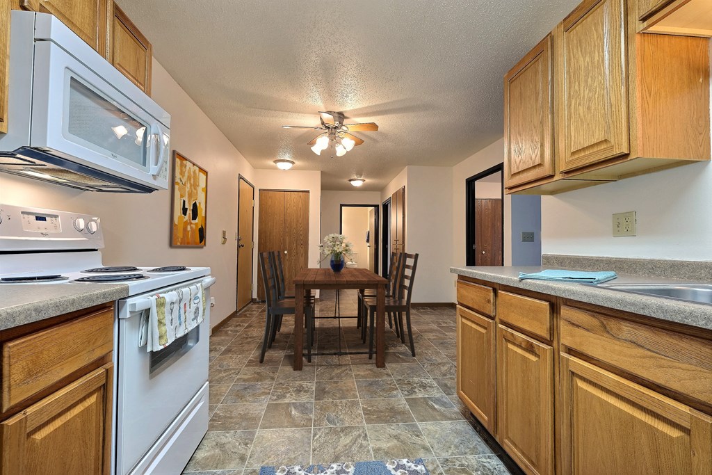 Fargo, ND Prairiewood Courts Apartments. A kitchen with white appliances and wooden cabinets. The dining room and table are in the background with four chairs and a light/ fan above it.