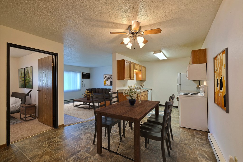 Fargo, ND Prairiewood Courts Apartments. A dining room and living room with a wooden table and four chairs and a ceiling fan. the living room is in the background.