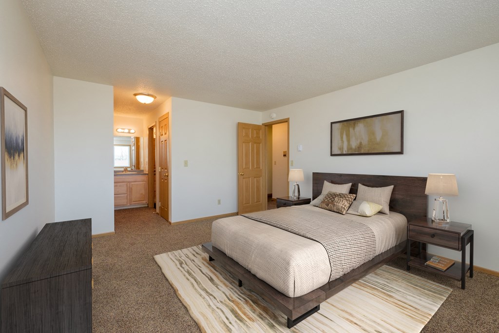 Fargo, ND Windgate Apartments. a master bedroom with a large bed and dresser. A bathroom is in the background