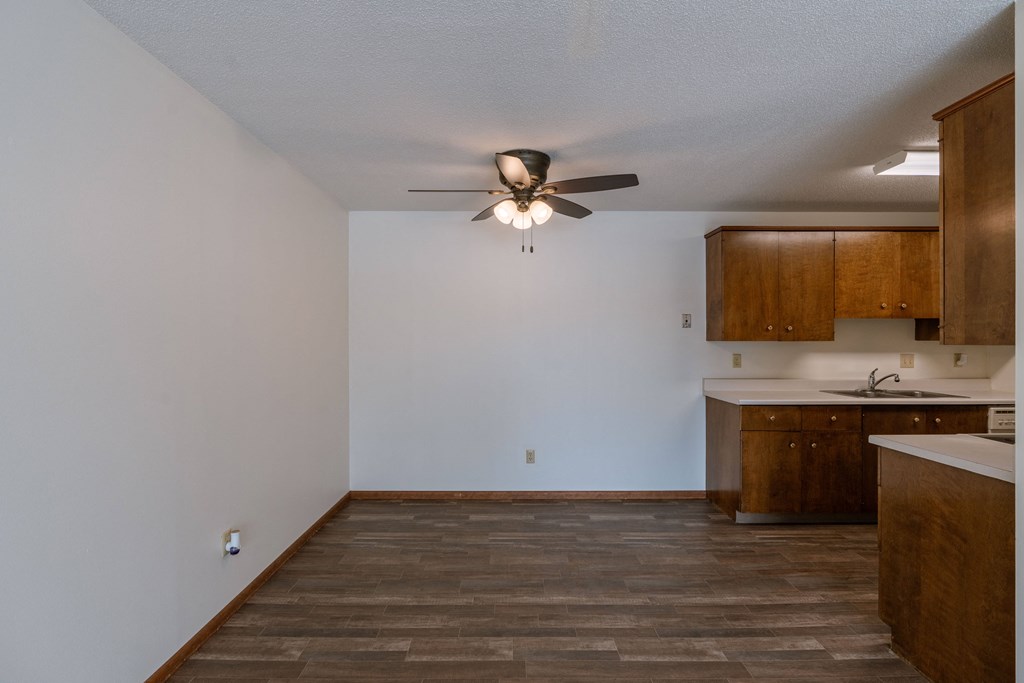 an empty living room with a ceiling fan and a kitchen. Fargo, ND Windsor Apartments