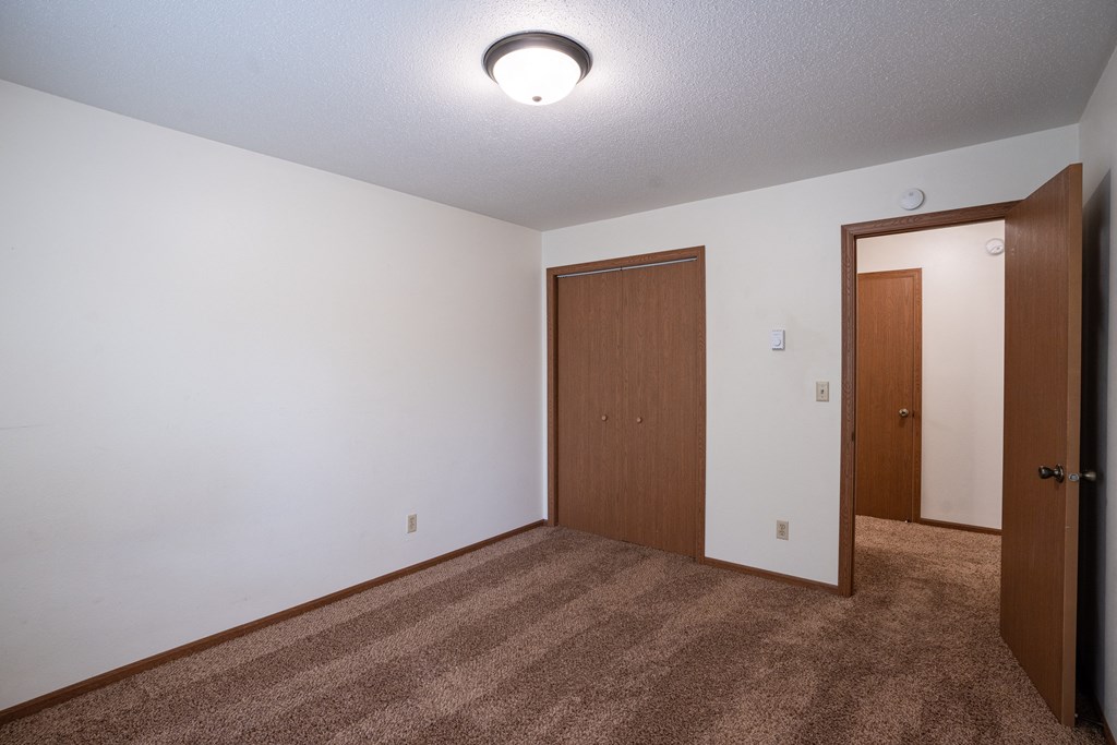 an empty living room with carpet and a door to a bedroom. Fargo, ND Monticello Apartments