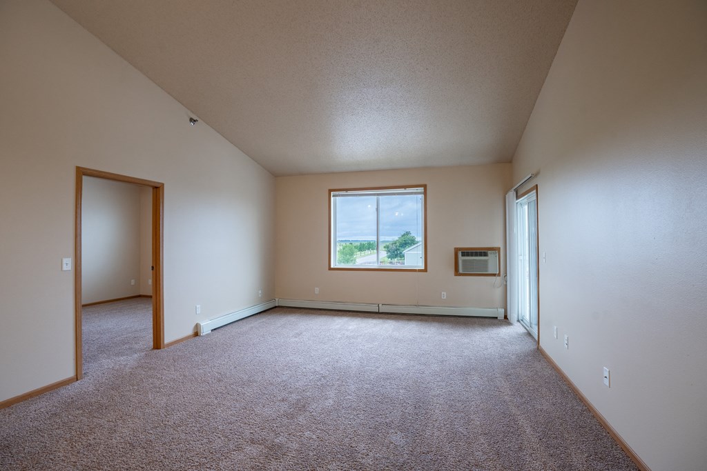 an empty living room with carpet and a window. Fargo, ND West Lake Apartments