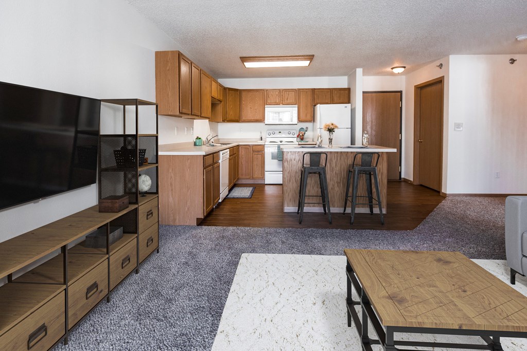 Grand Forks, ND Autumn Ridge. A living room with a tv, coffee table, and couch with a kitchen with bar stools at the island in the background.