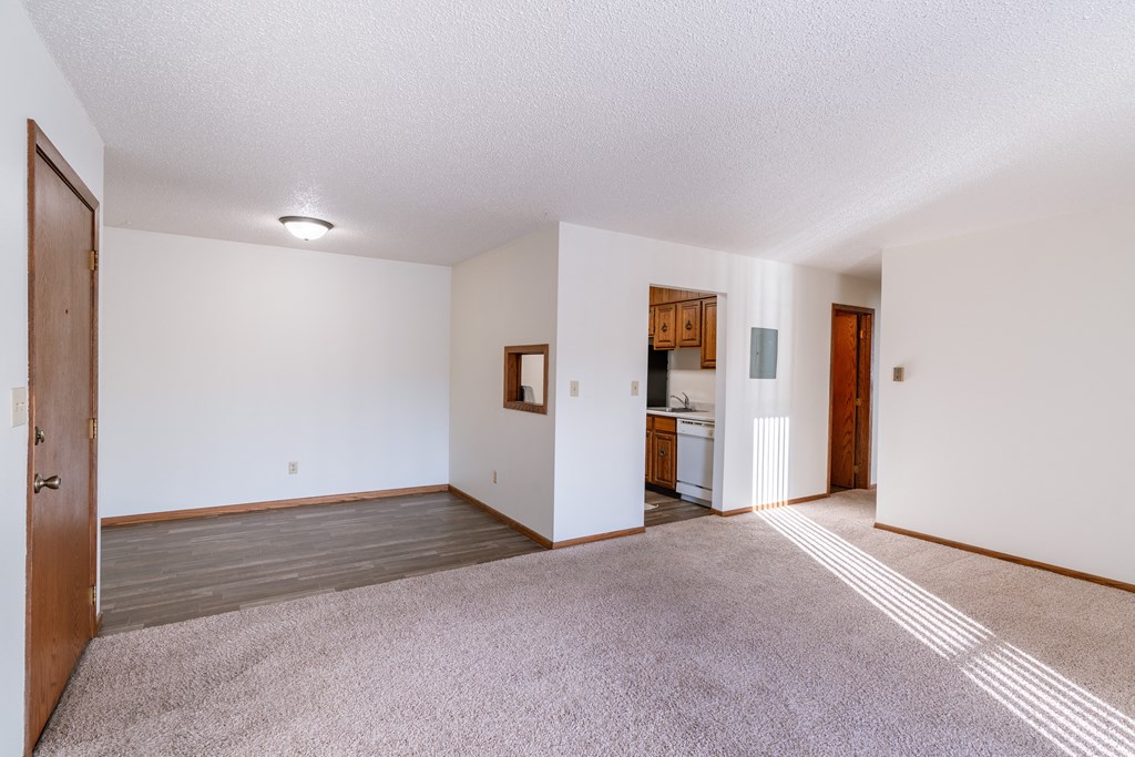 an empty living room with white walls and carpet and a kitchen. Grand Forks, ND Cedarwood Apartments