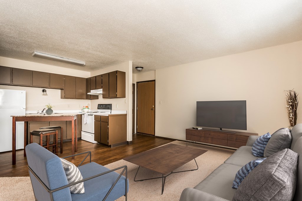 Grand Forks, ND Riverwood Apartments. A living room with a couch, chair, coffee table, and tv with the kitchen in the background.