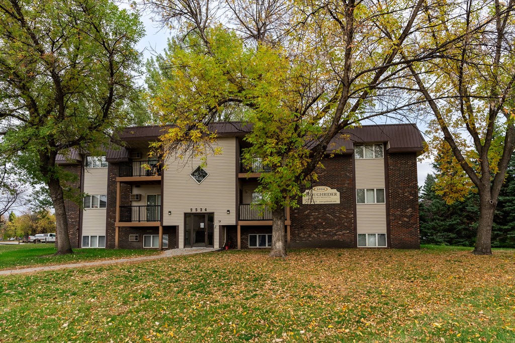 Grand Forks, ND Roughrider Apartment | Exterior Grand Forks, ND Roughrider Apartments. The exterior of a three level apartment building with a sidewalk leading to the front door.