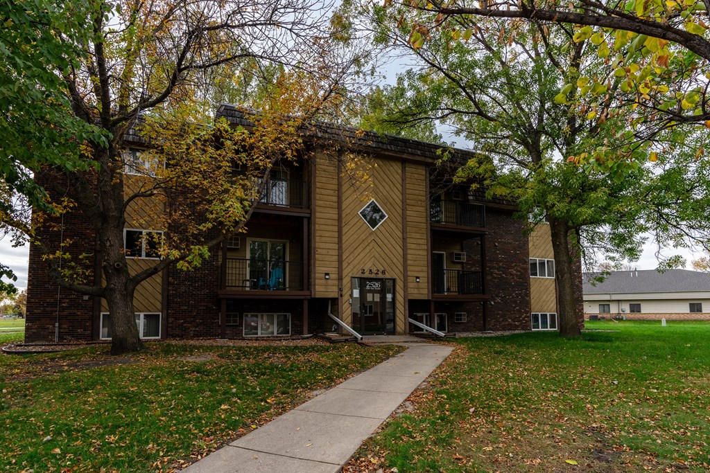 Grand Forks, ND Roughrider Apartment | Exterior Grand Forks, ND Roughrider Apartments. The exterior of a three level apartment building with a sidewalk leading to the front door.
