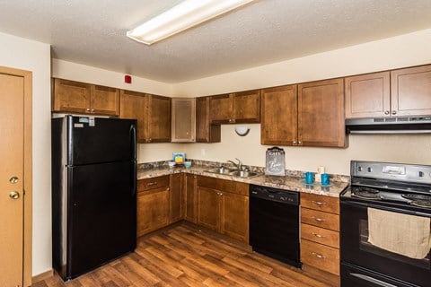 Grand Forks, ND Stanford Court Apartments. A kitchen with black appliances and a living room in the background a kitchen with wooden cabinets and black appliances