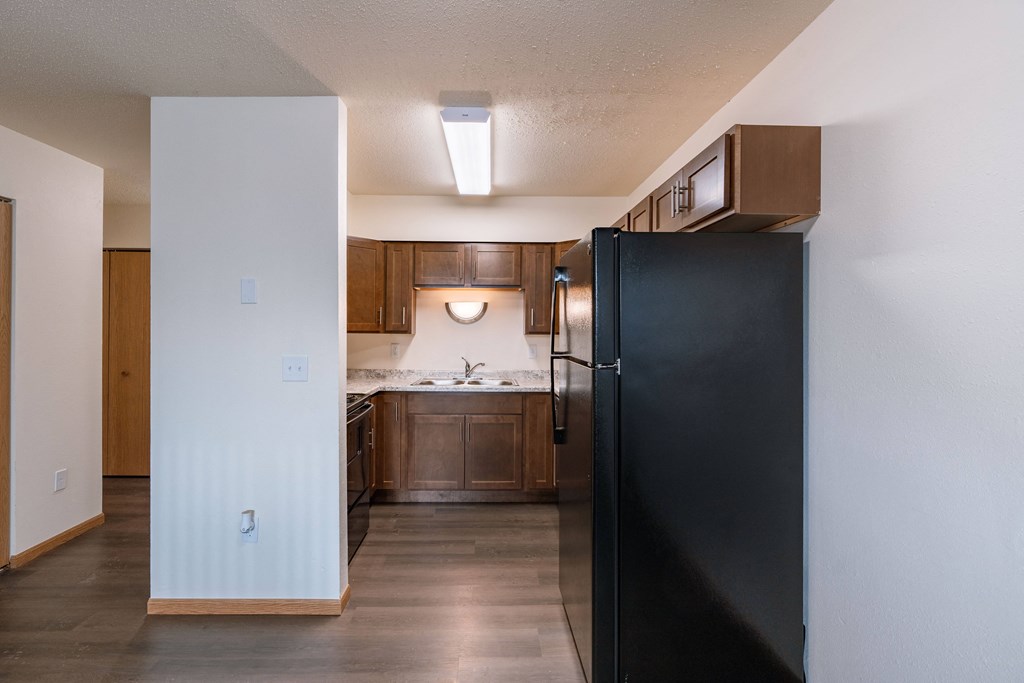 an empty kitchen with a black refrigerator and wooden cabinets. Moorhead, MN South Park Apartments