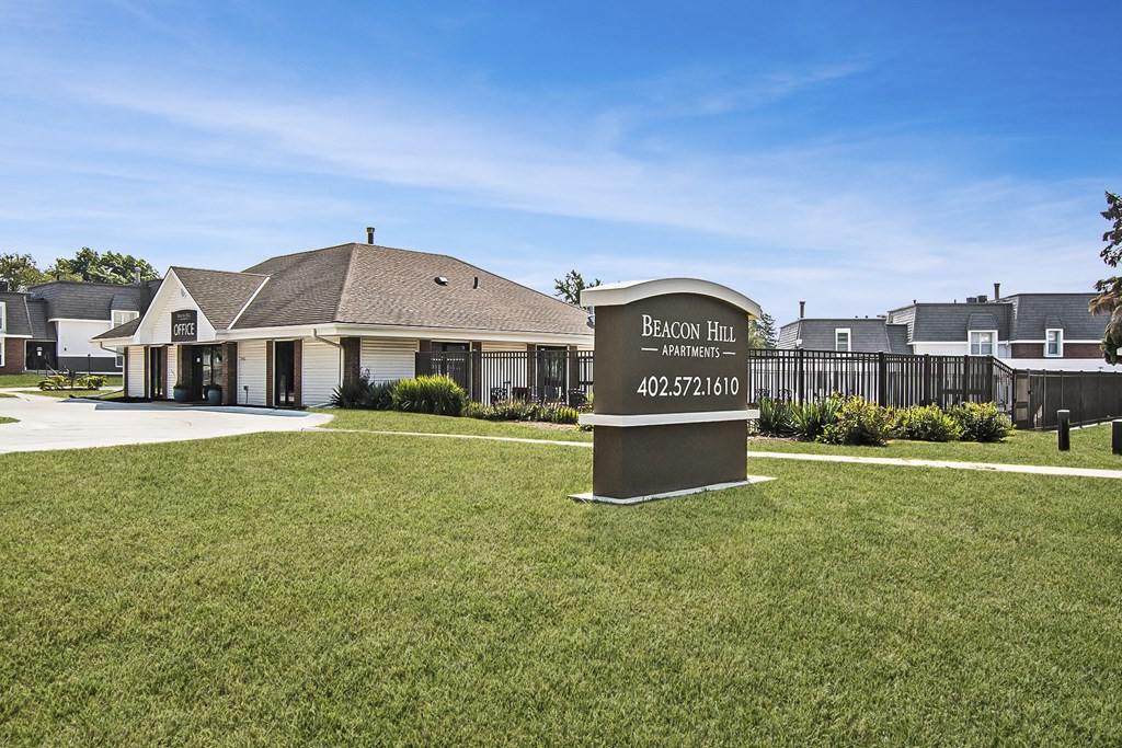 an exterior of an apartment office with a sign and green grass out front. Omaha, NE Beacon Hill Apartments.