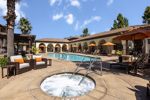 A hot tub sits in the middle of a patio with a pool and chairs.