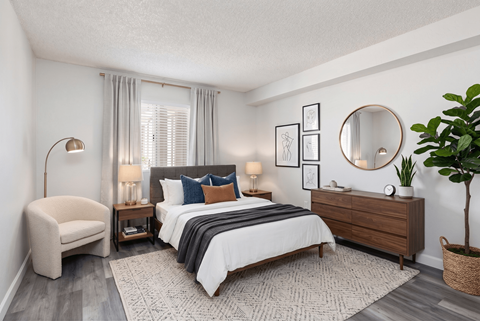 A bedroom with a large bed, a chair, a side table with lamps, a dresser, and a potted plant.