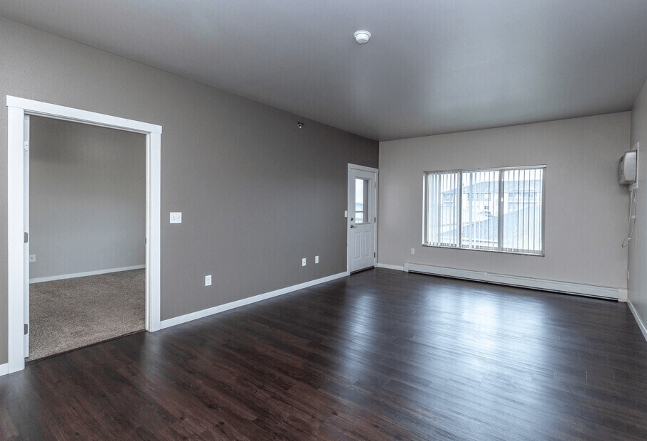 A large empty room with wood flooring and a window with blinds.
