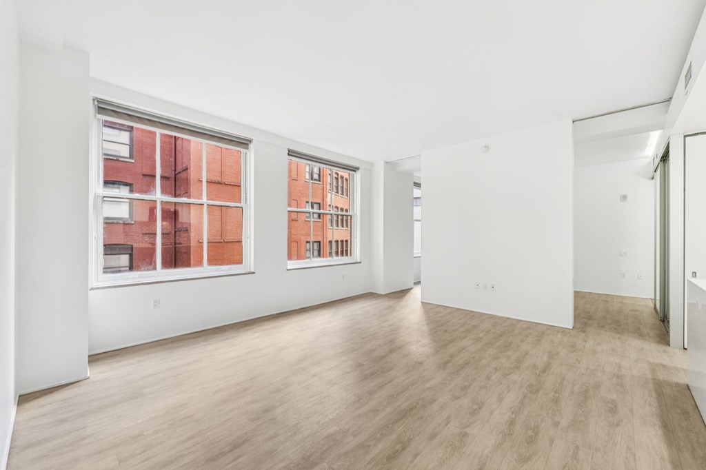 A room with white walls and wooden flooring with a window showing a red brick building outside.