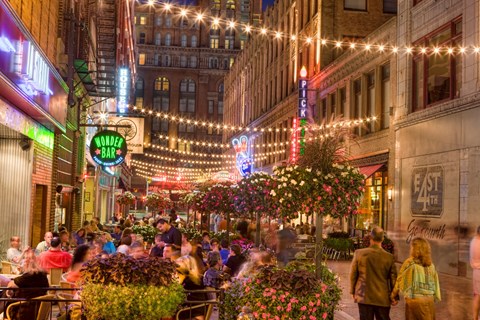 A busy street scene with people dining at tables and walking around.