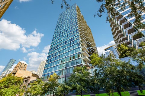 A tall building with many windows stands in front of other buildings and trees.
