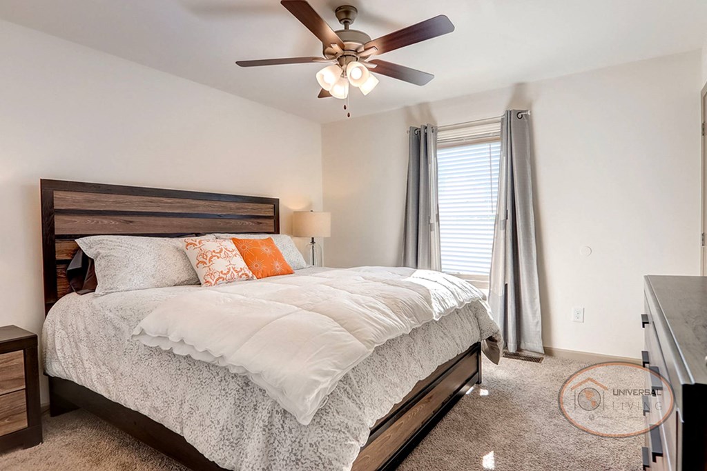 A bedroom with a large bed, large window, carpet, ceiling fan, and white walls.
