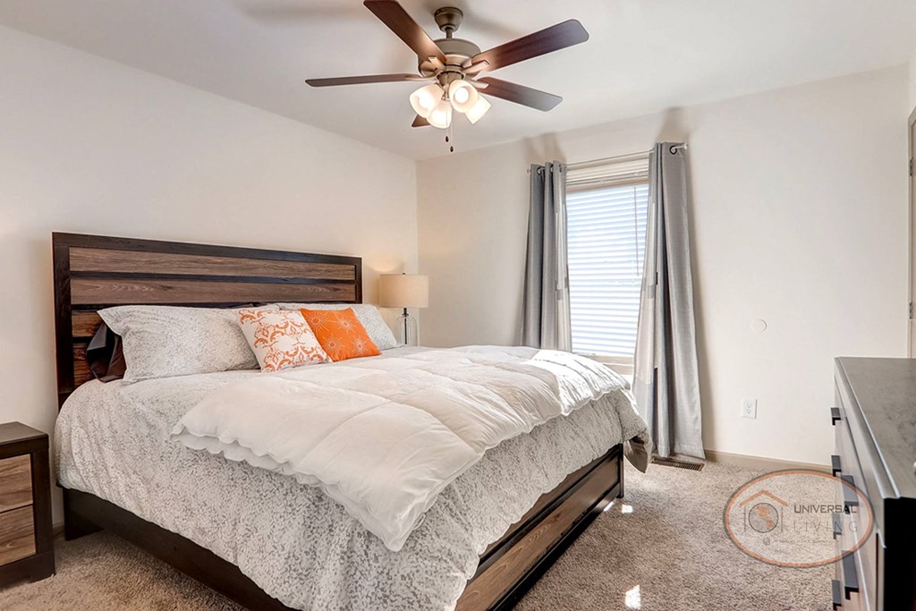 A bedroom with a large bed, white walls, carpet, and  ample natural light.