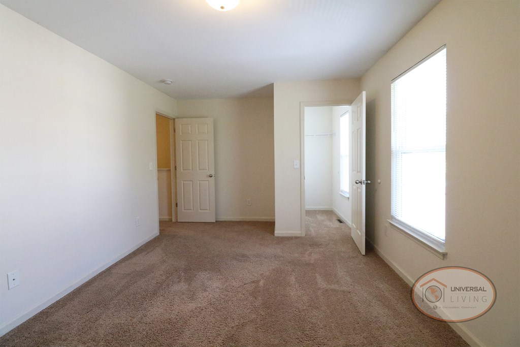 An empty bedroom with walk in closet, bright natural light, tan carpet, and white walls.