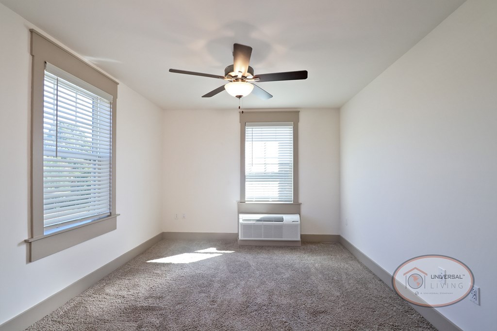 A bedroom with white walls, tan carpet, two large windows and a ceiling fan.