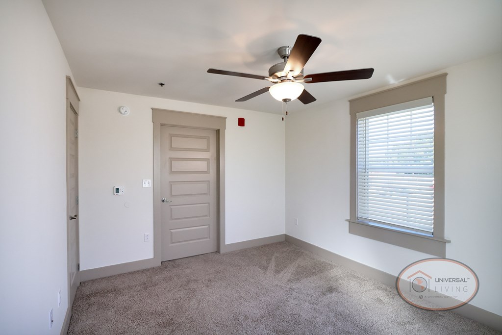 A bedroom with carpet, white walls, a ceiling fan, large window and a closet.