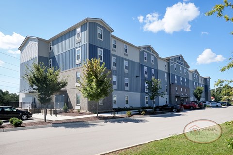 The front of a large blue and grey apartment building surrounded by lush green trees.