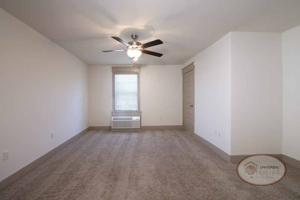 The living area with white walls, carpet, a large window and a ceiling fan.