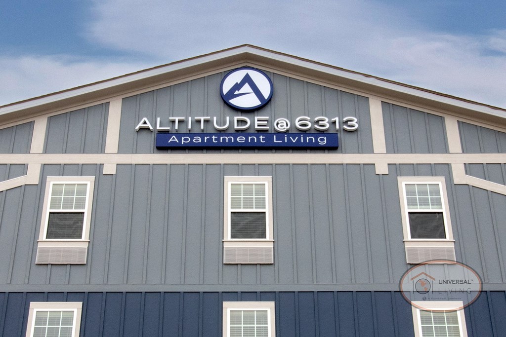 The sign on the top of the blue and grey apartment building that reads "Altitude @ 6313 Apartment Living."