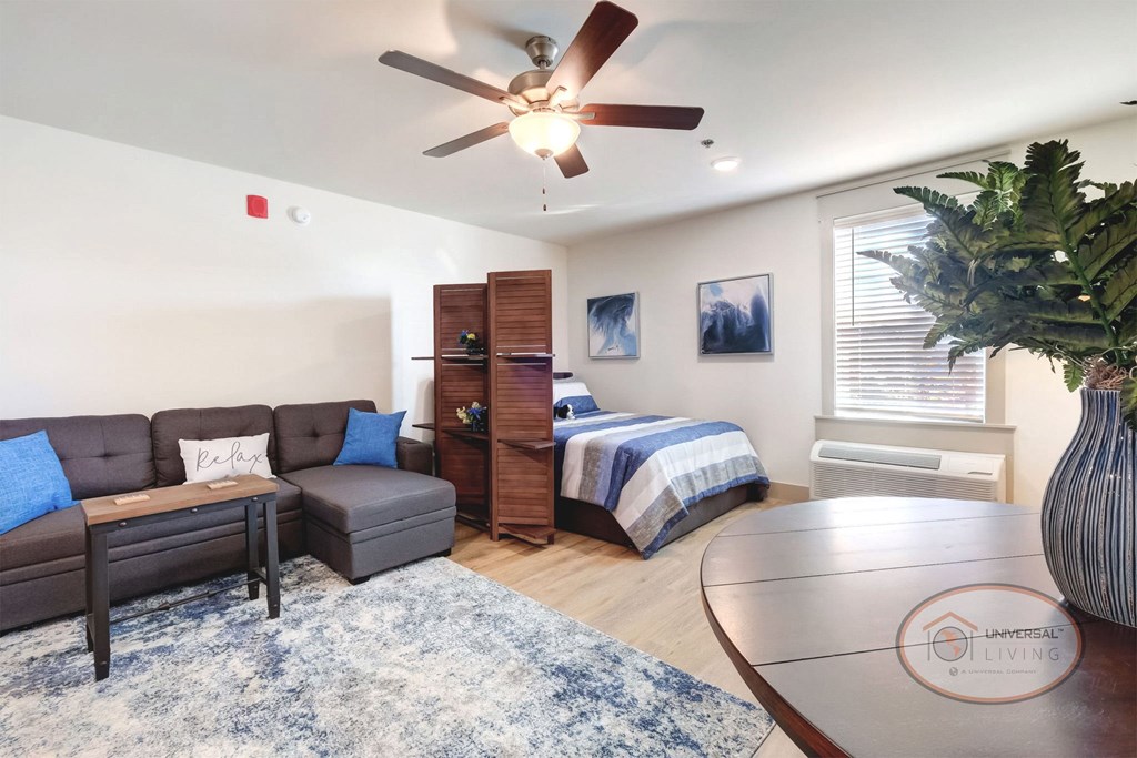 The living and sleeping area in a studio apartment with white walls, vinyl flooring, and a ceiling fan.
