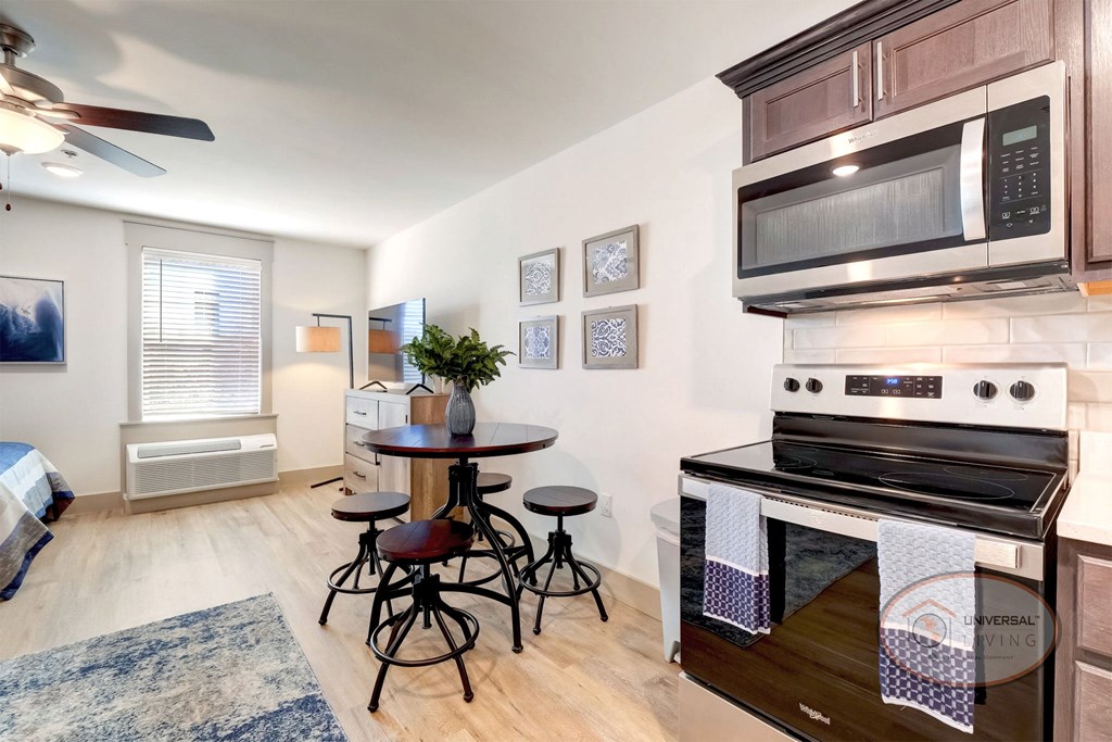 A studio apartment kitchen with stainless steel appliances, a small table, and a large window.