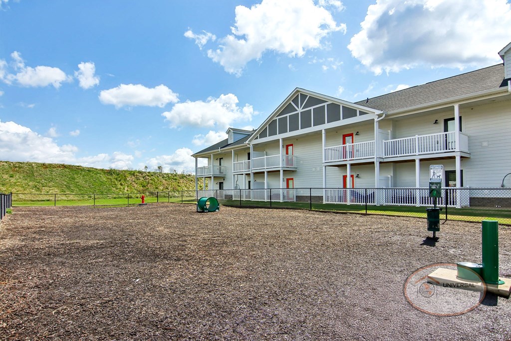 A fenced in dog park with a water fountain and obstacles for your dog with an apartment building in the background.