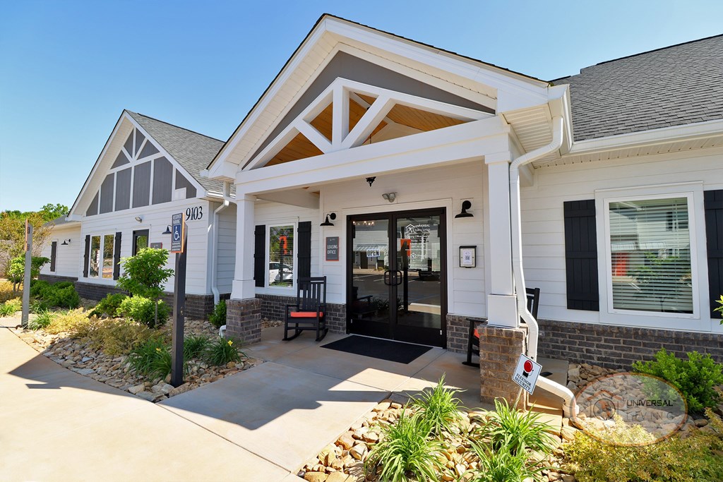 The front of the grey and white clubhouse with green plants and rocking chairs on the porch.