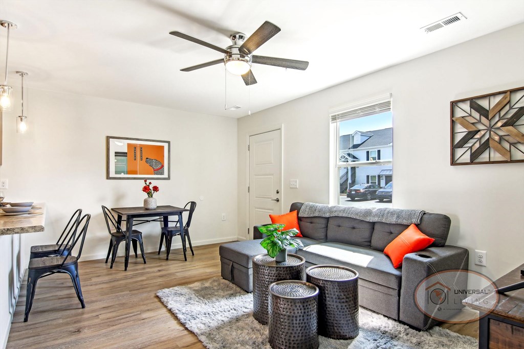 An open window lets light into a white living room with a grey couch, a dining table with chairs, and a ceiling fan.